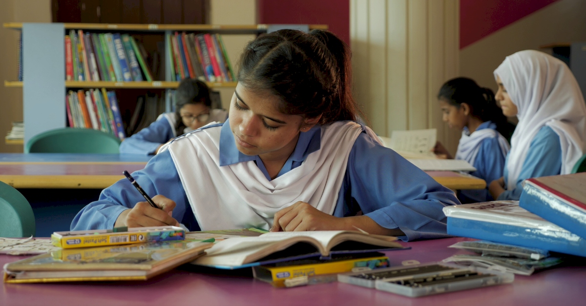 A student in a blue and white uniform looks down while drawing at her desk. Other female students sit behind her, reading.