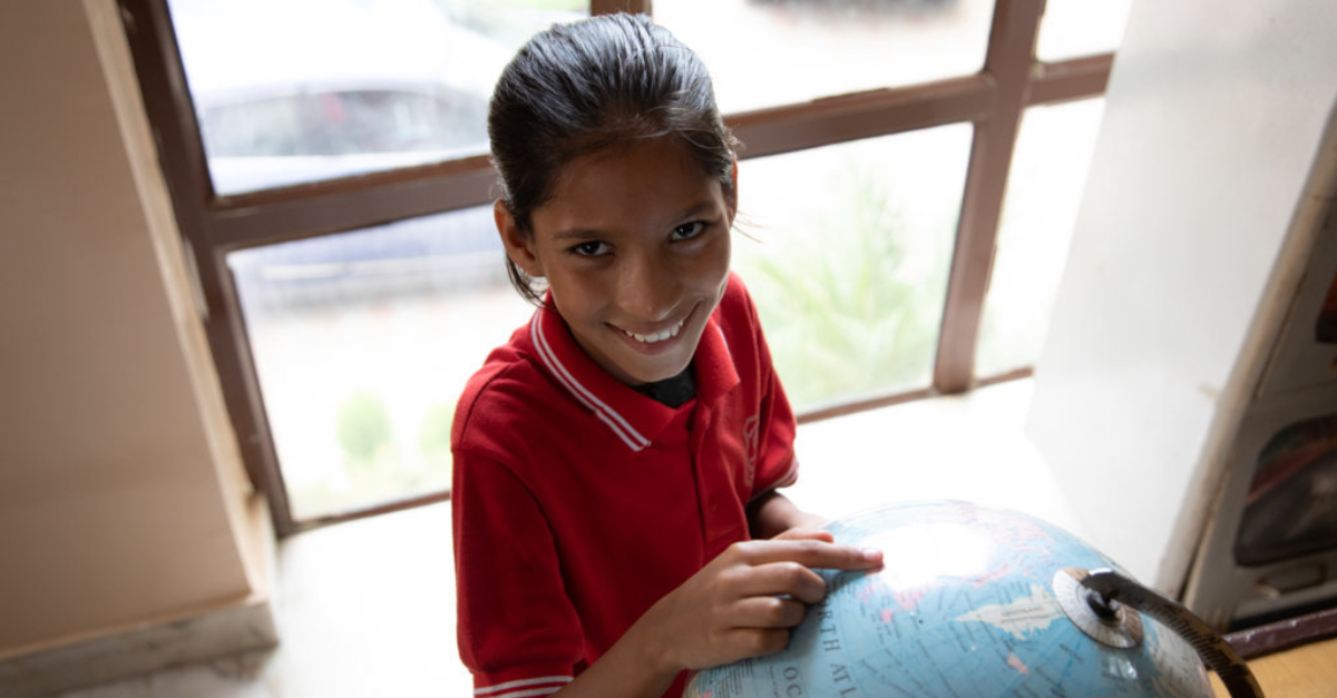 A young girl smiles at the camera and points to a country on a globe.